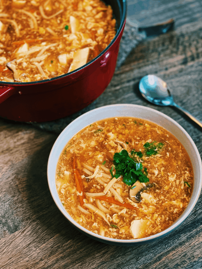 Bowl of steaming Hot and Sour Soup, with visible slices of tofu, carrots, and shiitake mushrooms, garnished with chopped spring onions, offering a tantalizing blend of spicy and tangy flavors.