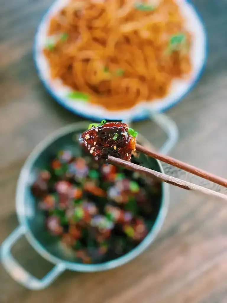 Chopsticks holding up a piece of sweet and sour pork spare ribs with a bigger pot of the dish as well as stir fried noodles in the background