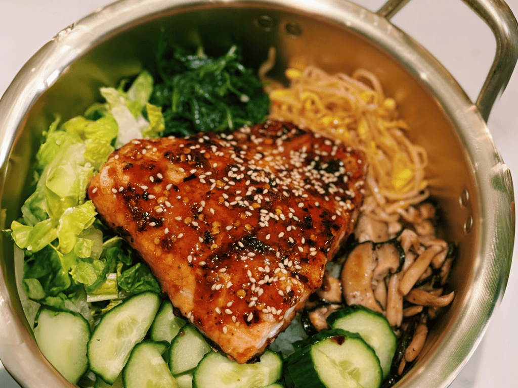 Succulent spicy honey glazed salmon over bibimbap, surrounded by an array of colorful Korean side dishes. The salmon's rich glaze shines atop the mixed rice bowl, complemented by vibrant side dishes like kimchi and seasoned vegetables. This image captures the essence of a wholesome, flavor-packed Korean meal, appealing to food lovers seeking a blend of sweet, spicy, and savory tastes.