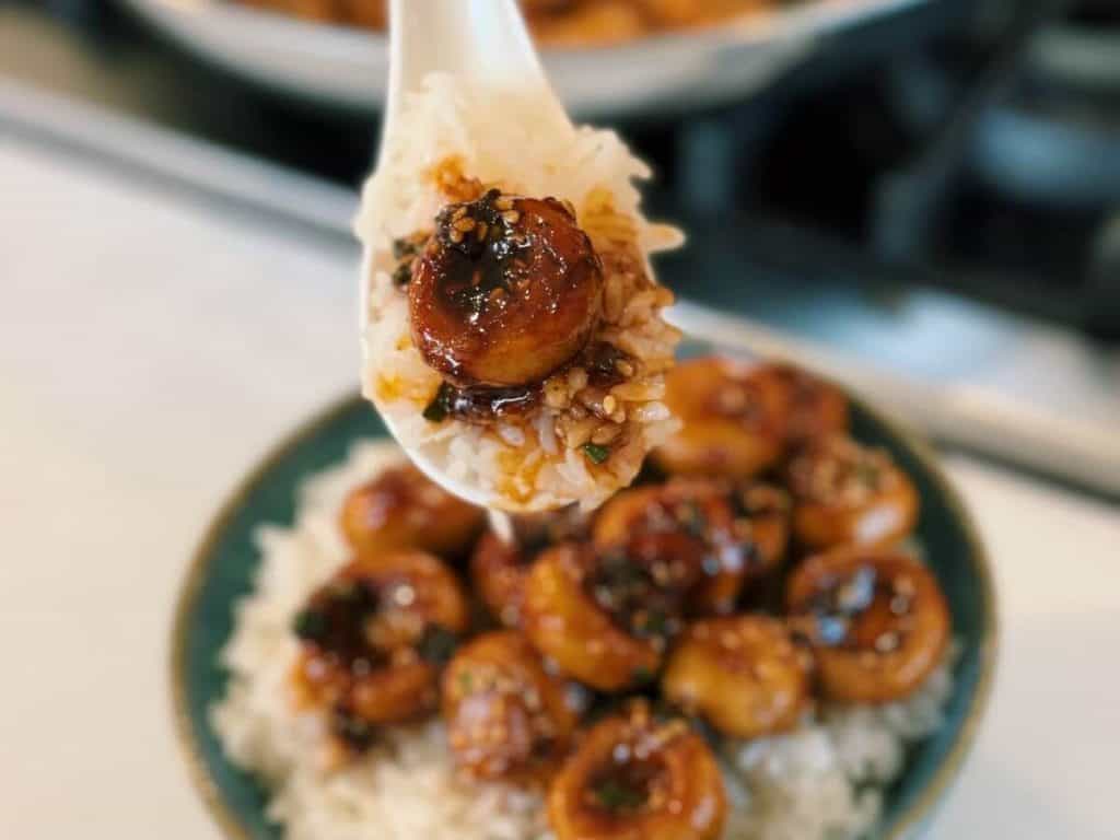 A spoonful of mushrooms from a bowl of Sweet and sour mushrooms over a bowl of fluffy white rice, garnished with green onions and sesame seeds, ready to eat