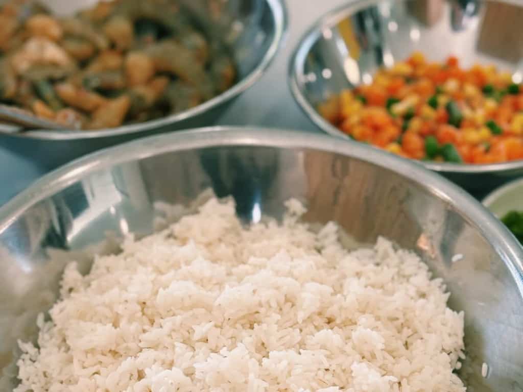 A showcase of ingredients for shrimp fried rice, including marinated shrimp, cold separated rice, and vegetables in metal bowls