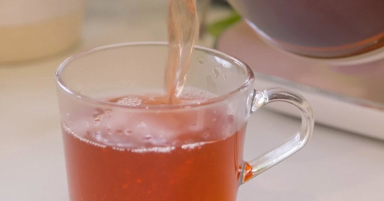 A cup of pinkish red Herbal Hibiscus Rose Beauty Tea poured from a kettle into a clear cup