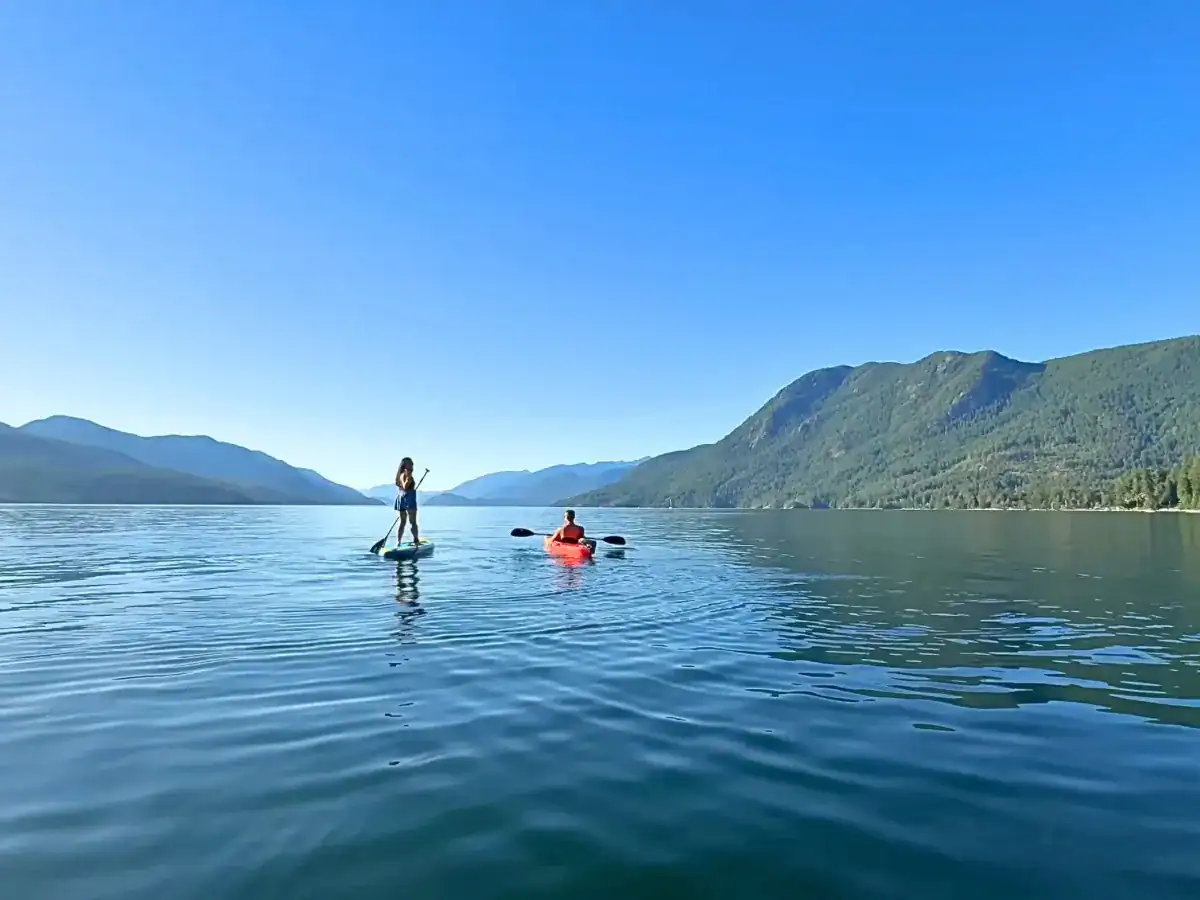 Serene lake with two people kayaking and paddleboarding amid mountains on a clear sunny day.