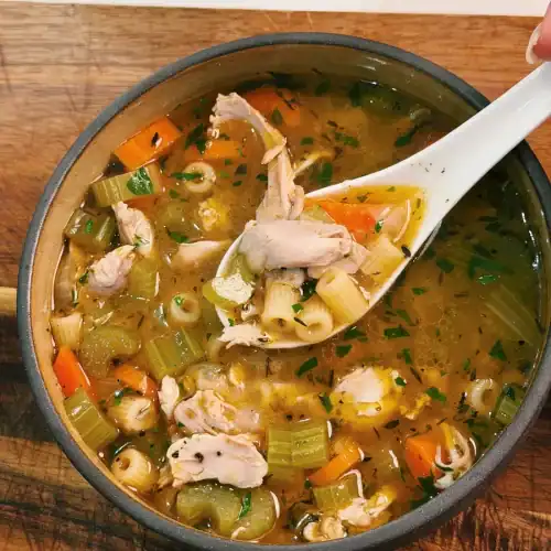 A bowl of chicken noodle soup with a scoop lifted to show ingredients, including chicken thigh, carrot, celery, and onions.