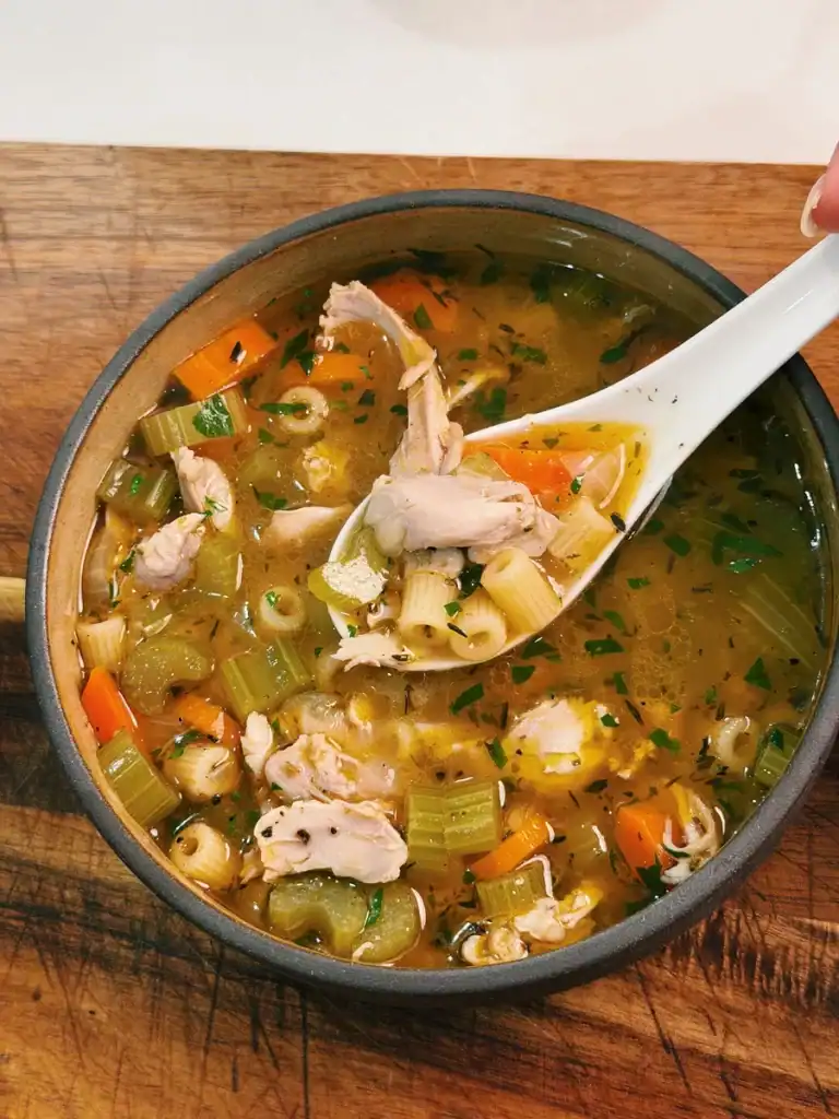 A bowl of chicken noodle soup with a scoop lifted to show ingredients, including chicken thigh, carrot, celery, and onions.