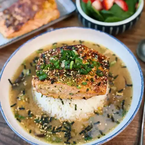 A bowl of Miso Salmon Brothy Rice with a serving of salmon and salad in the background showing a full meal