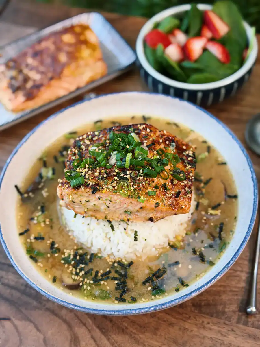 A bowl of Miso Salmon Brothy Rice with a serving of salmon and salad in the background showing a full meal