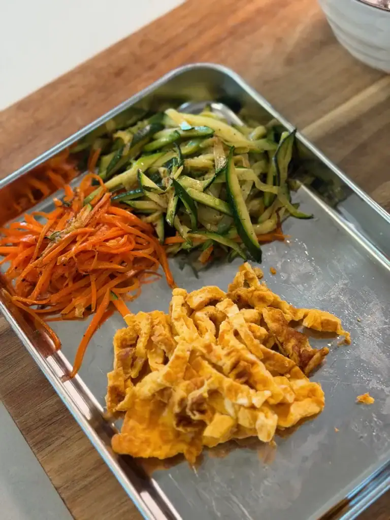 Sauteed vegetables like zucchini and carrots, and egg ribbons, as toppings for breakfast vegetable noodle soup