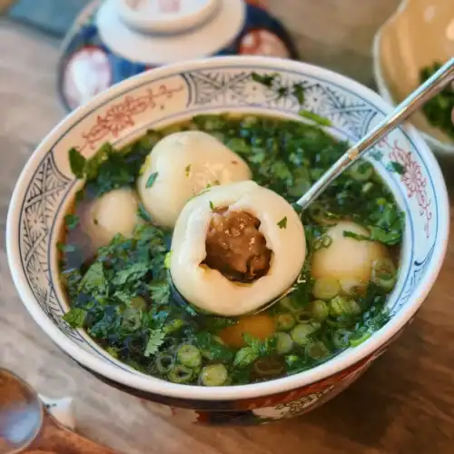 A bowl of savory Tang Yuan in light broth with a bite taken out of one showing the pork mushroom filling inside
